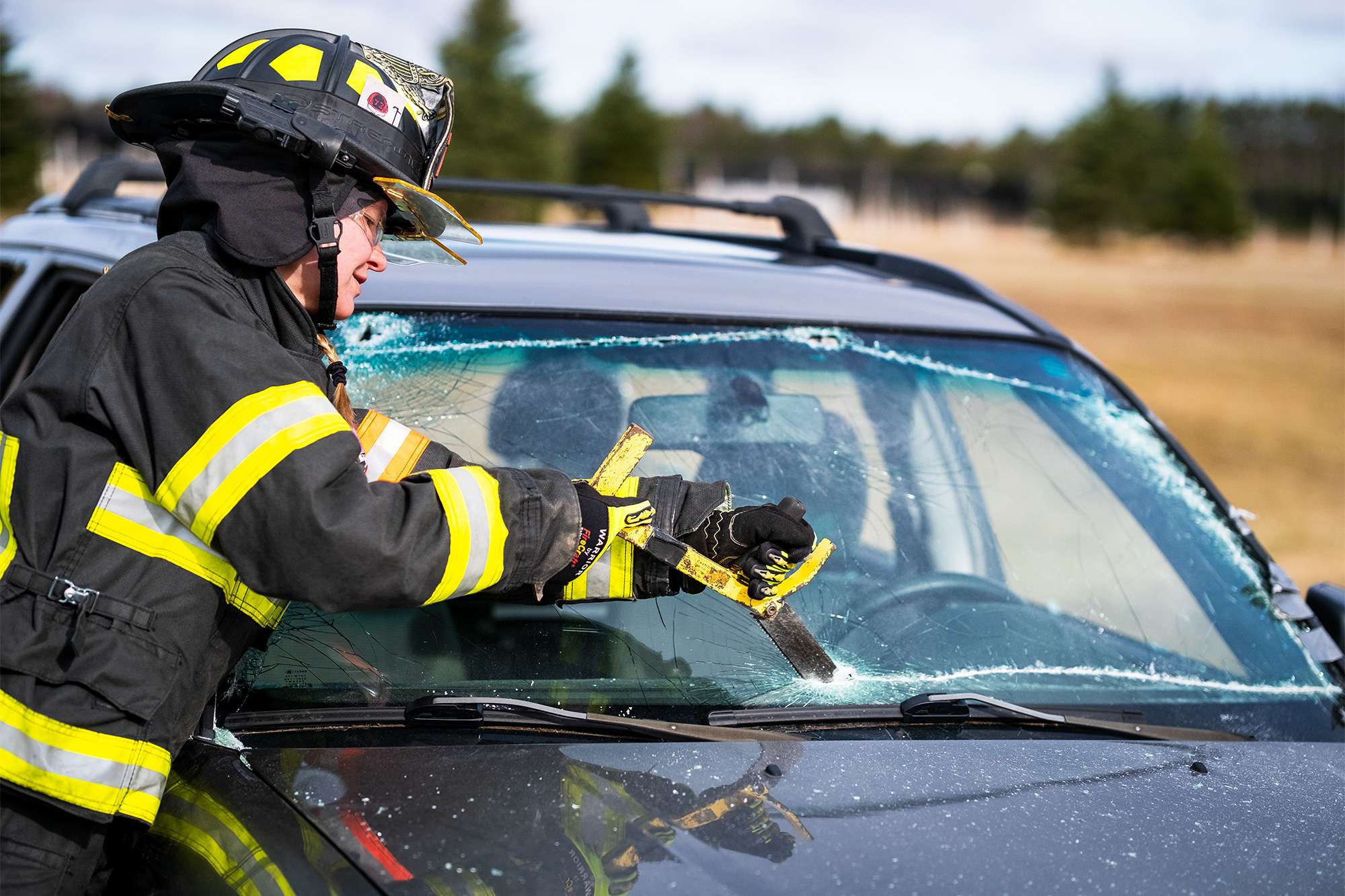 Firefighter in protective gear uses a tool to break a vehicle’s windshield during extrication training.