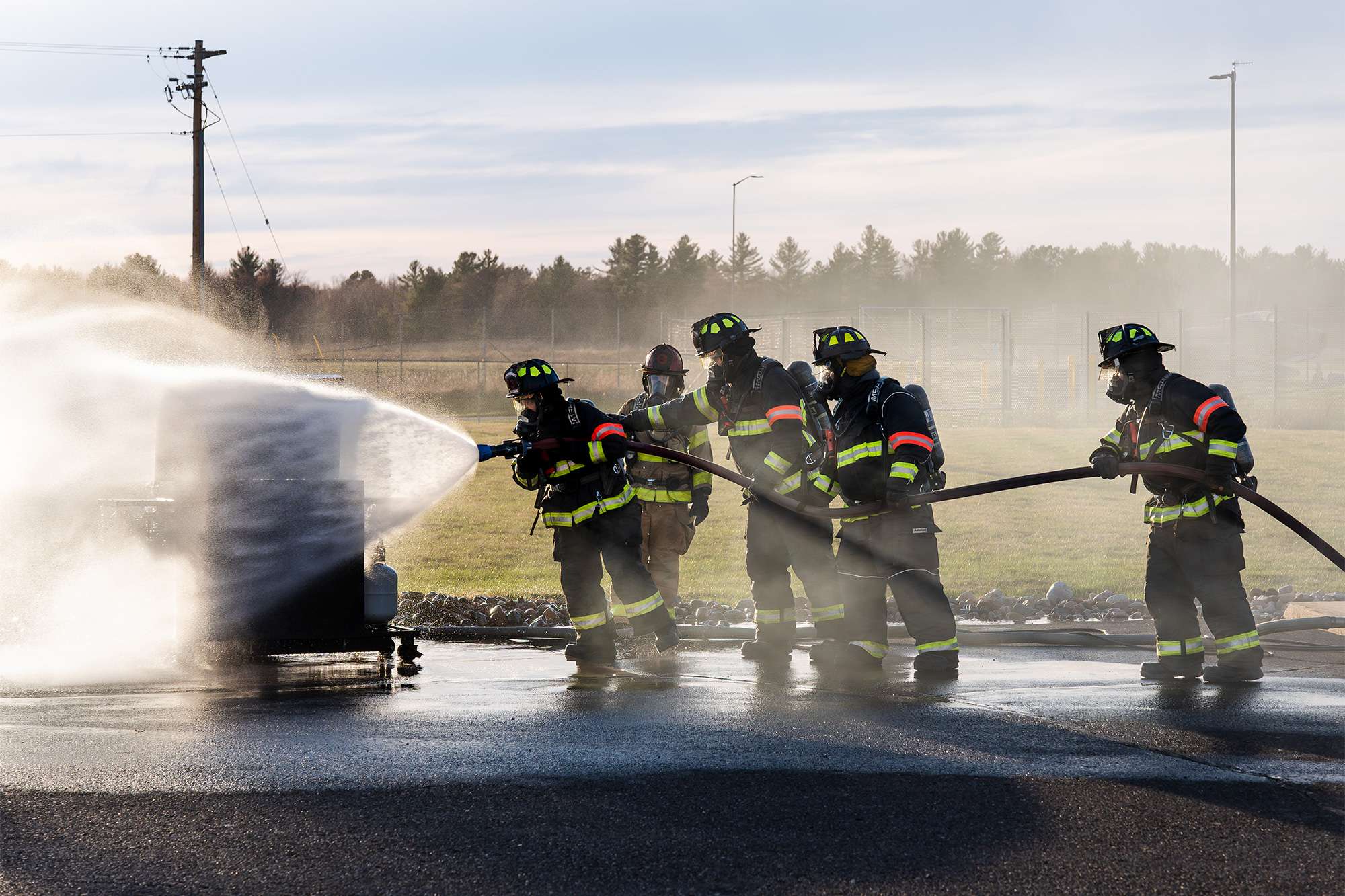 Firefighters in full gear work together to spray water onto a smoking metal unit during outdoor training.