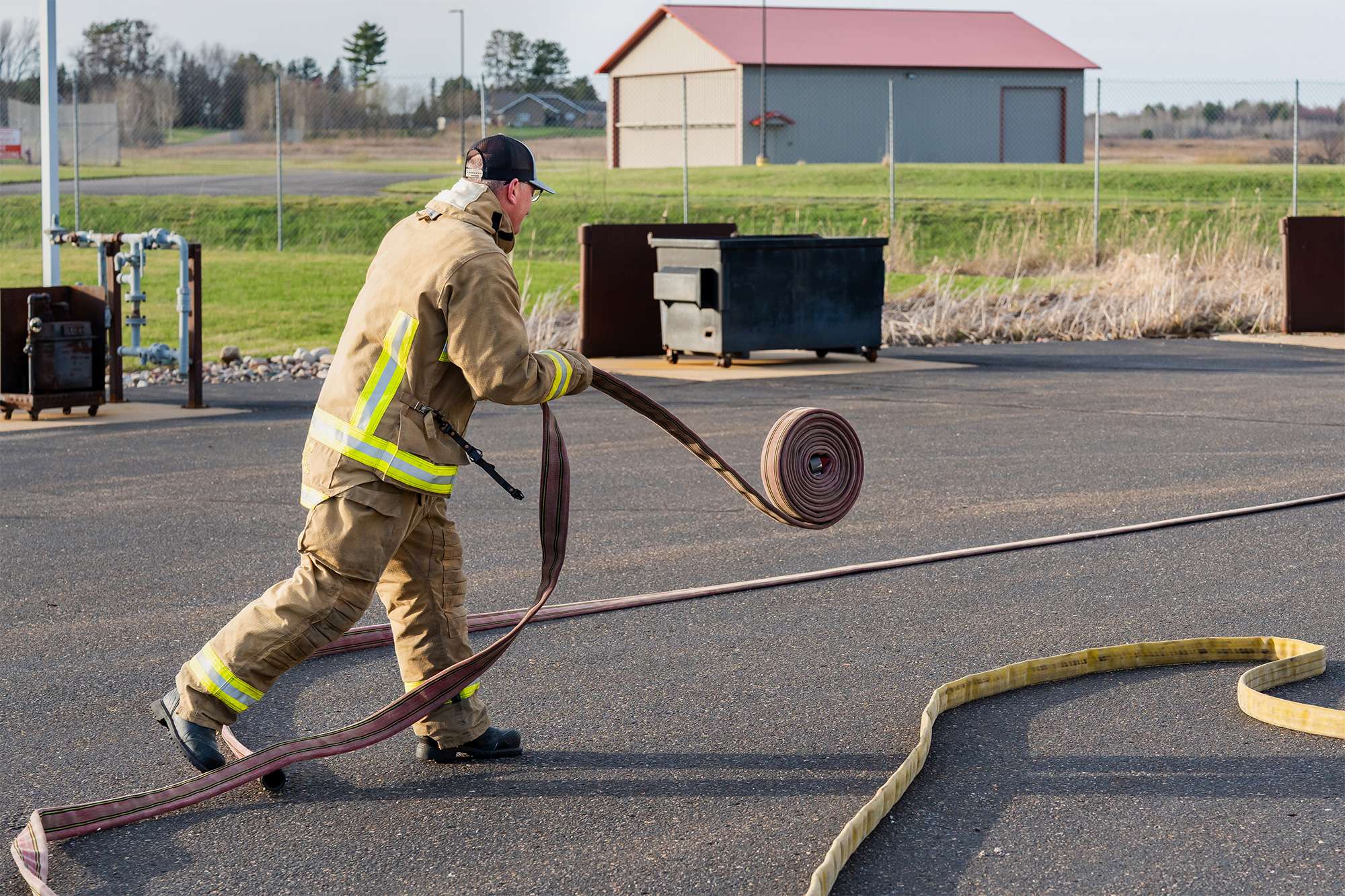 Firefighter in gear unrolls a hose on pavement during outdoor training.
