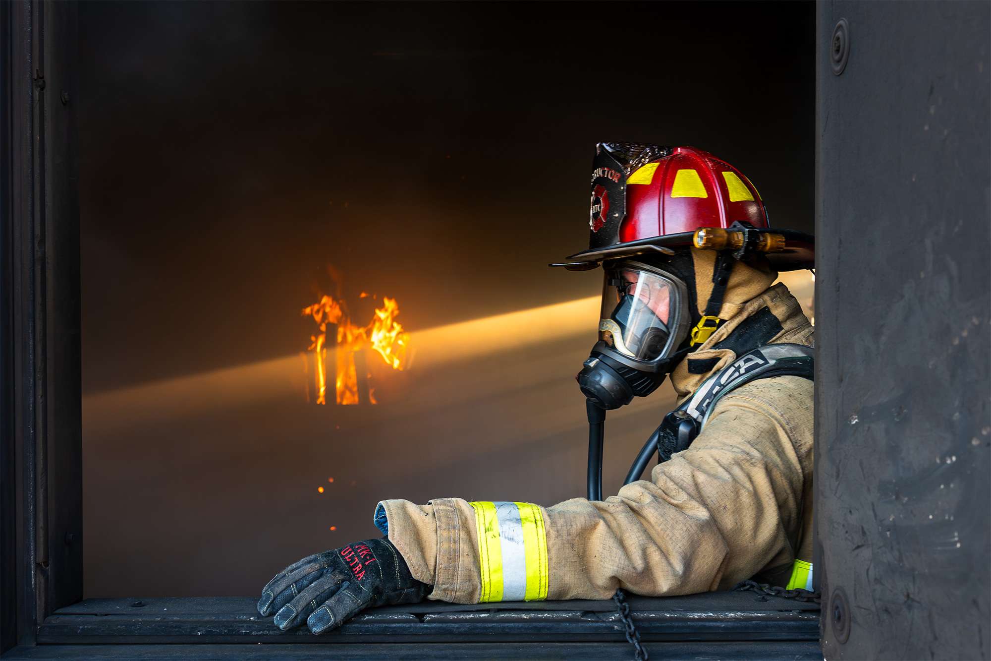 Firefighter in gear leans at a window as flames and a beam of light shine through smoke inside the training room.