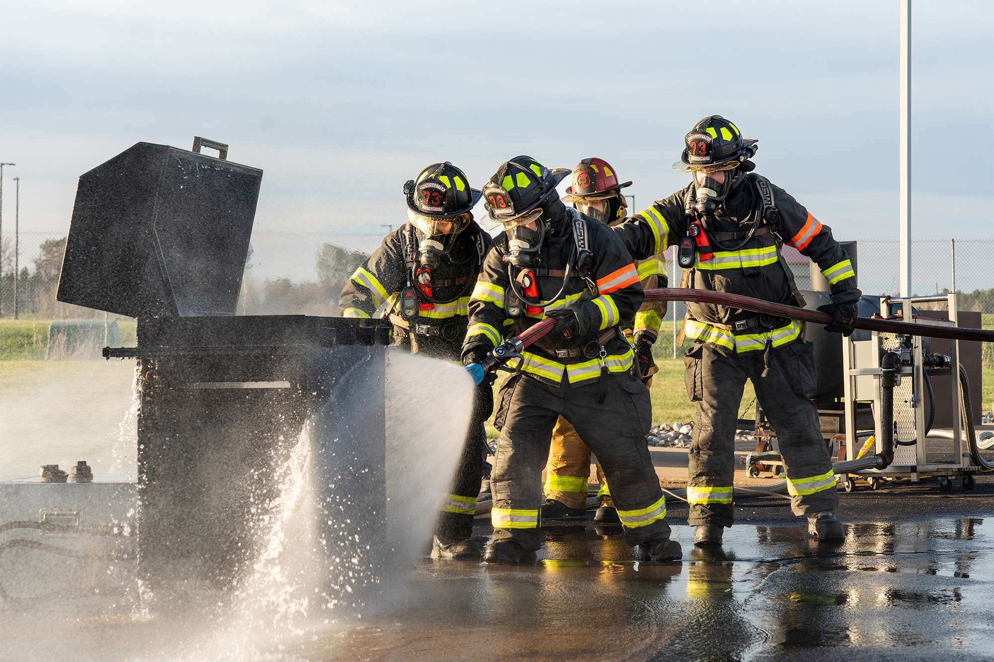 Firefighters in full gear use a hose to spray water onto a smoking metal container during training.
