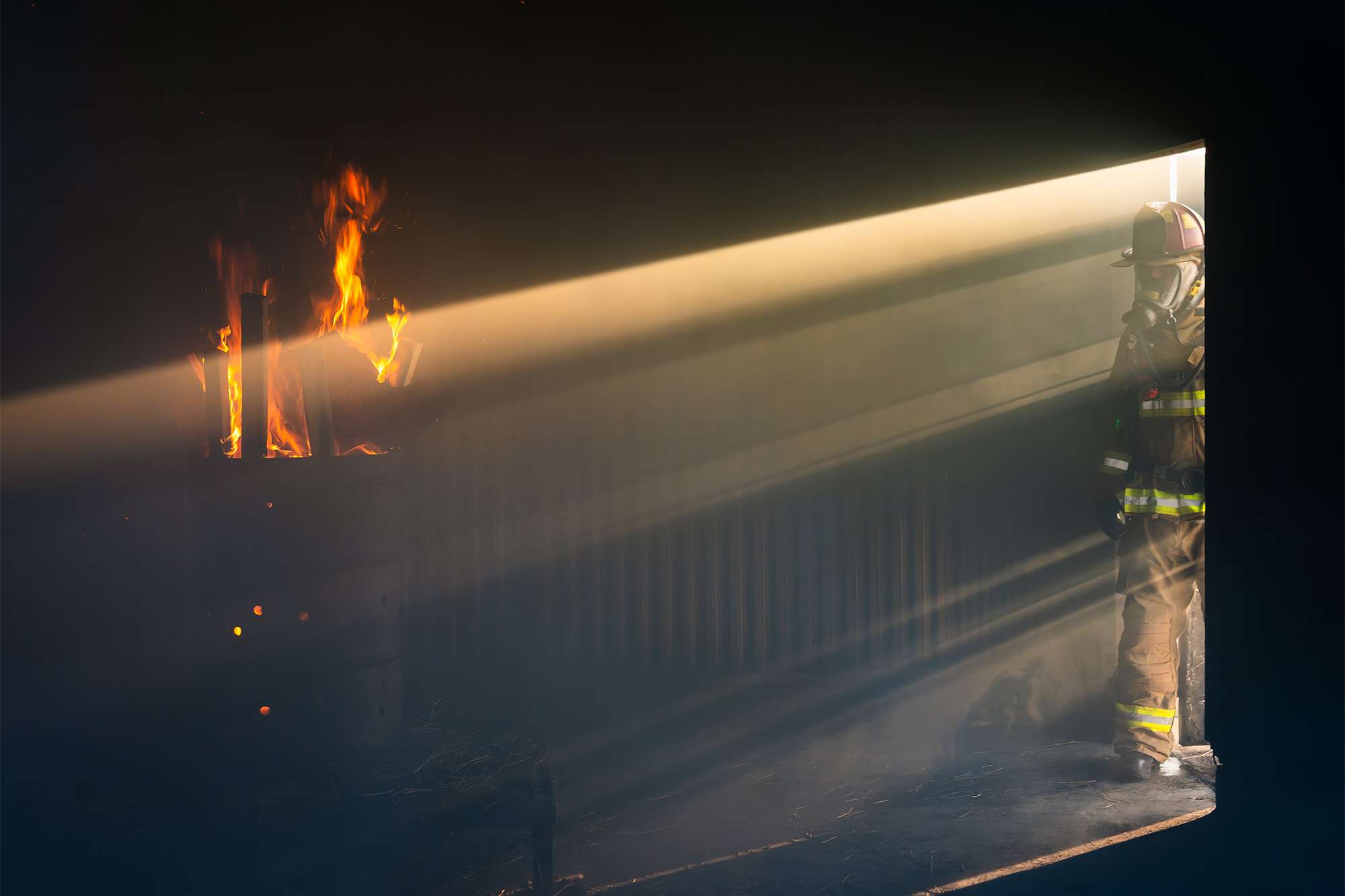 Firefighter standing in doorway as sunlight cuts through smoke toward a burning barrel inside a dark room.