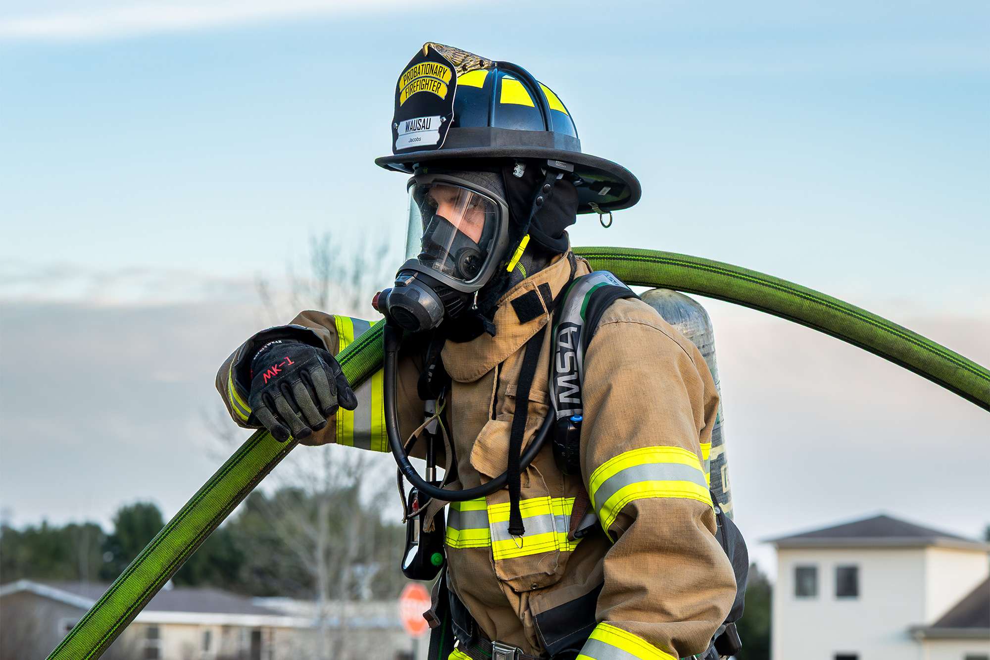 Firefighter in tan colored gear, helmet, and gas mask, with a green hose on his shoulder, looking left against a bright sky.