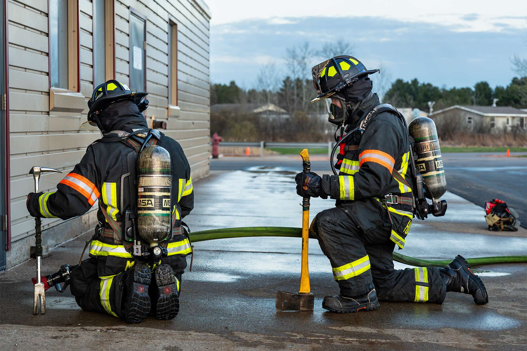Two firefighters in full gear kneel outside a building, preparing tools and hose during training.