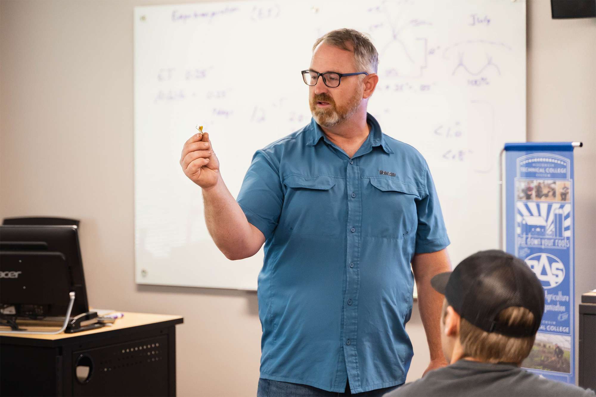 Instructor in blue shirt and glasses gestures with marker toward students, smiling; whiteboard with scribbles.