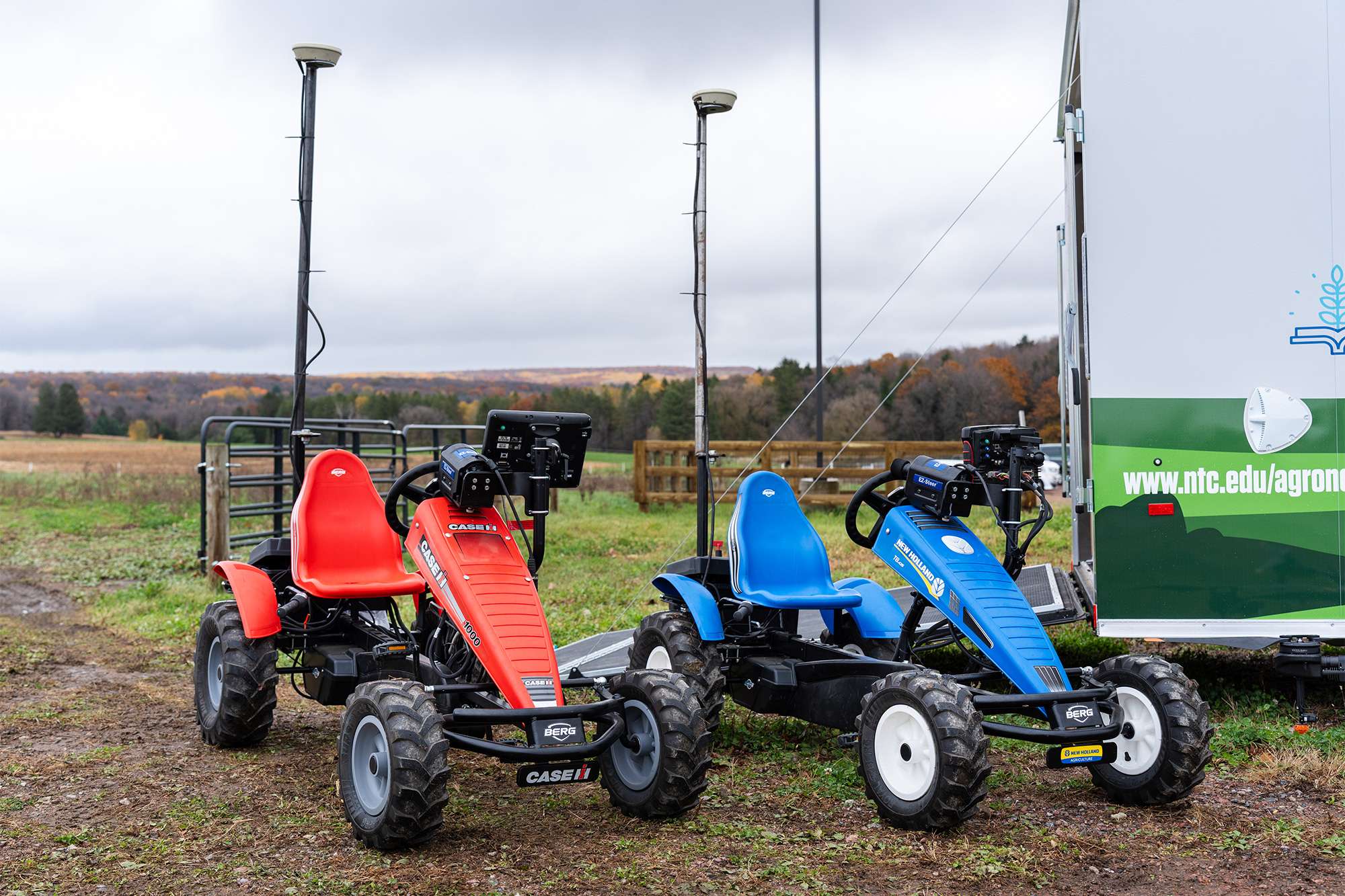 Two small off-road go-karts — one red, one blue — parked side-by-side on muddy grass near a green trailer.