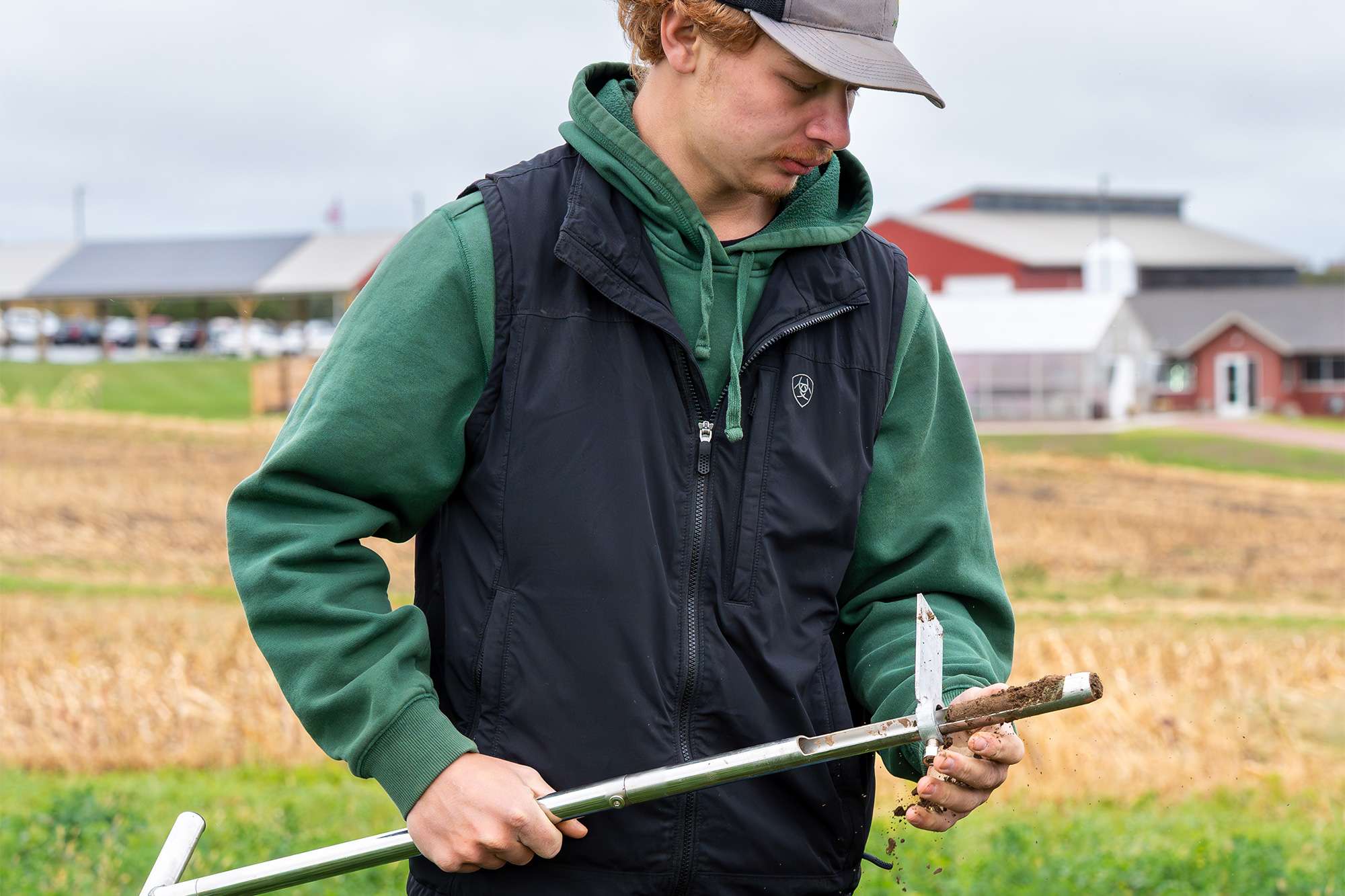 Young man in green hoodie and black vest outdoors in a harvested field, wearing a cap while adjusting a soil probe.
