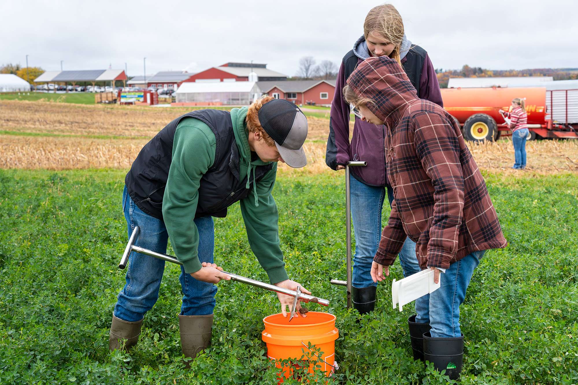 Three people in jackets inspect plants in a green field beside an orange bucket, one bending to collect samples.