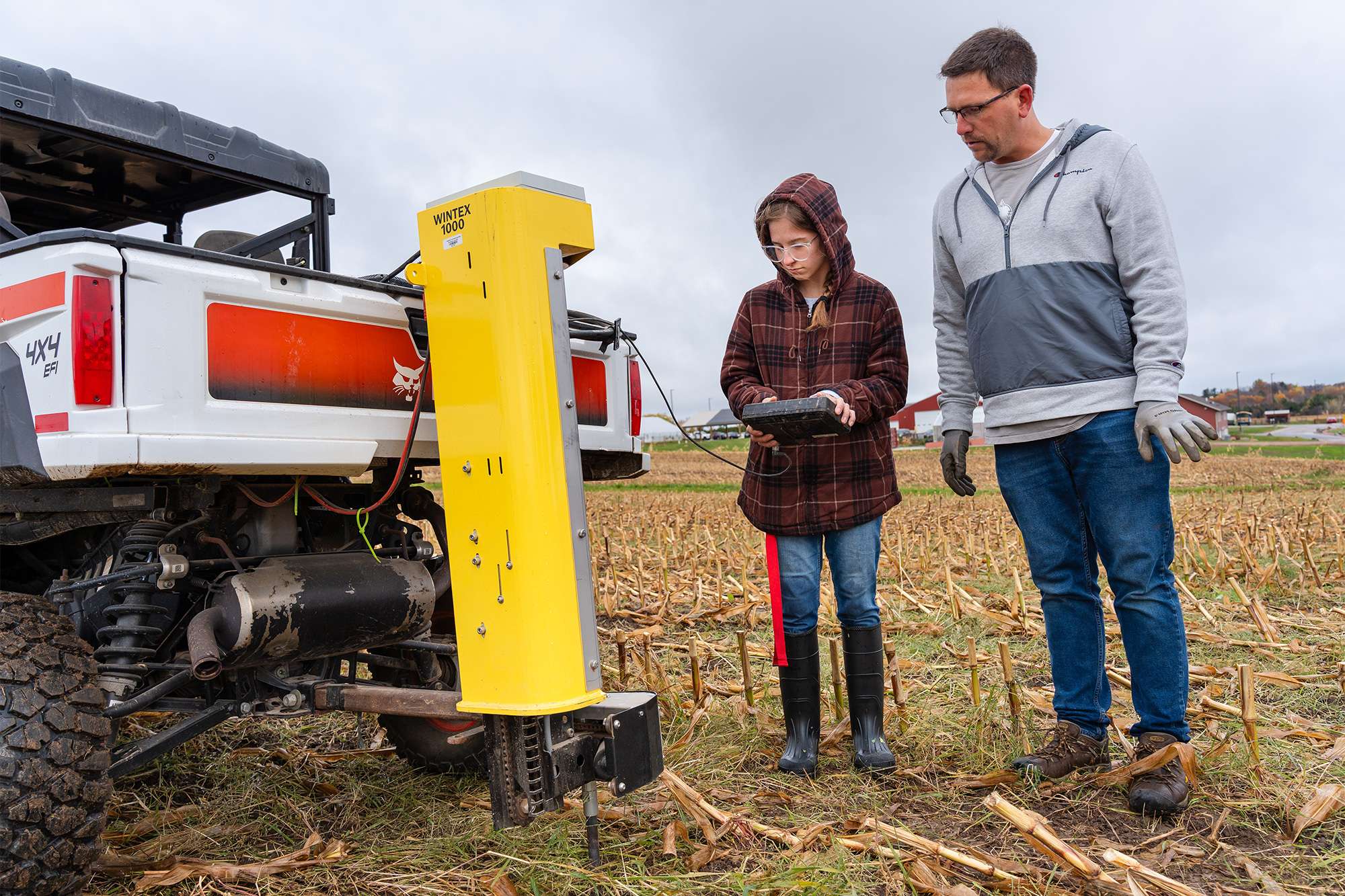 Man and teenage girl consult a tablet beside a white pickup with a yellow agricultural machine mounted in a cornfield.
