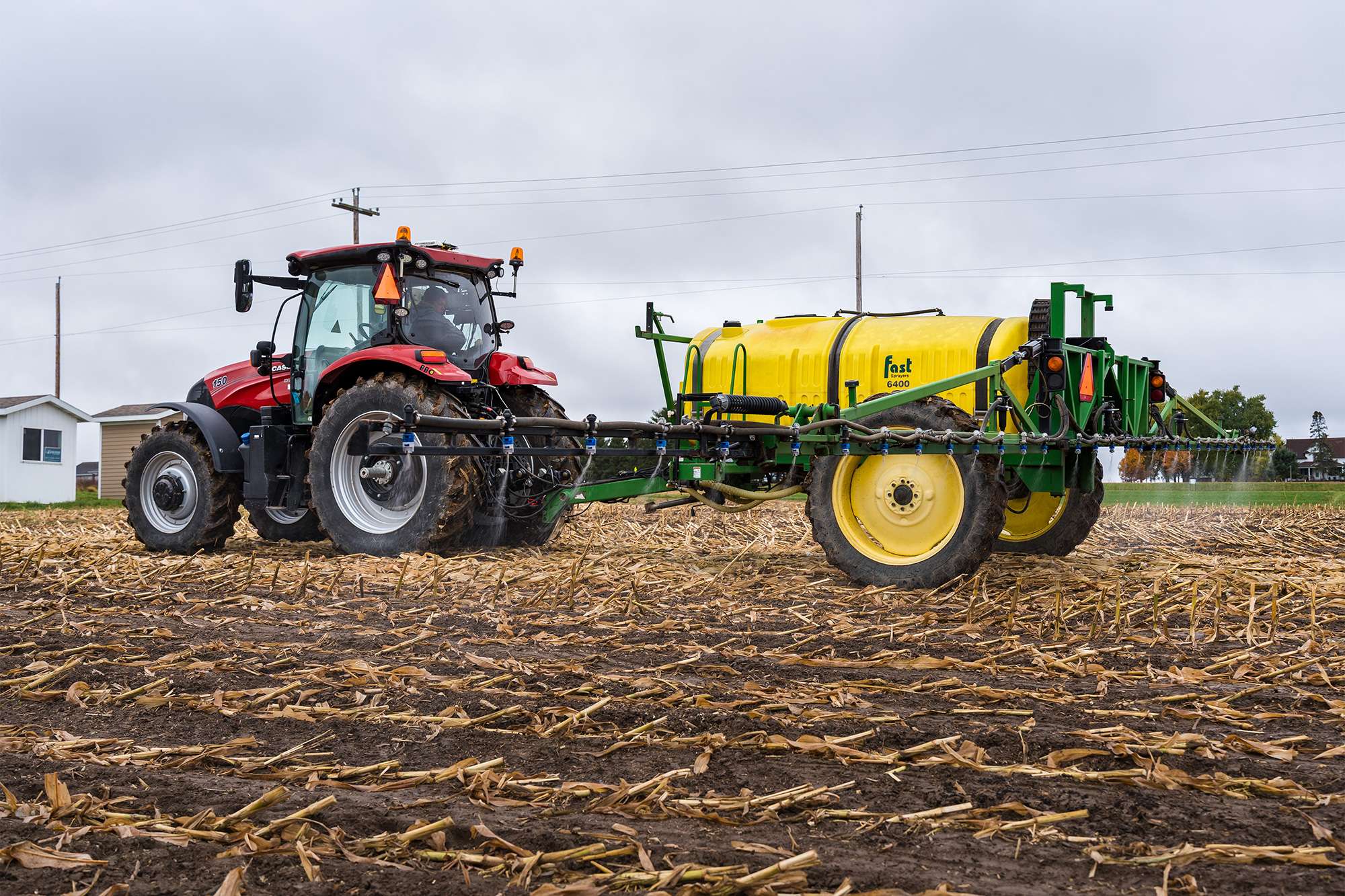 Red tractor towing a large yellow tank sprayer across a muddy, harvested field under an overcast sky.