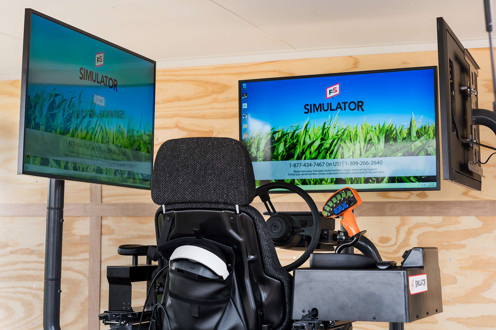 Black simulator seat and control console with joystick and throttle facing three large monitors showing a grassy landscape.