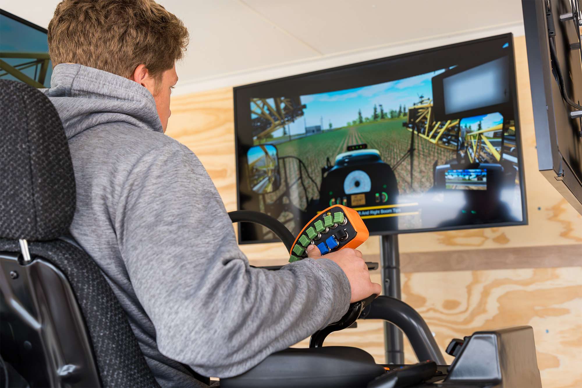 Person using a driving simulator, holding a controller while viewing a first-person driving scene on a large monitor.